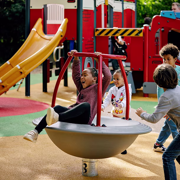 Children play on a spinning bowl ride in a colourful playground with a large red climbing structure and yellow slide in the background.