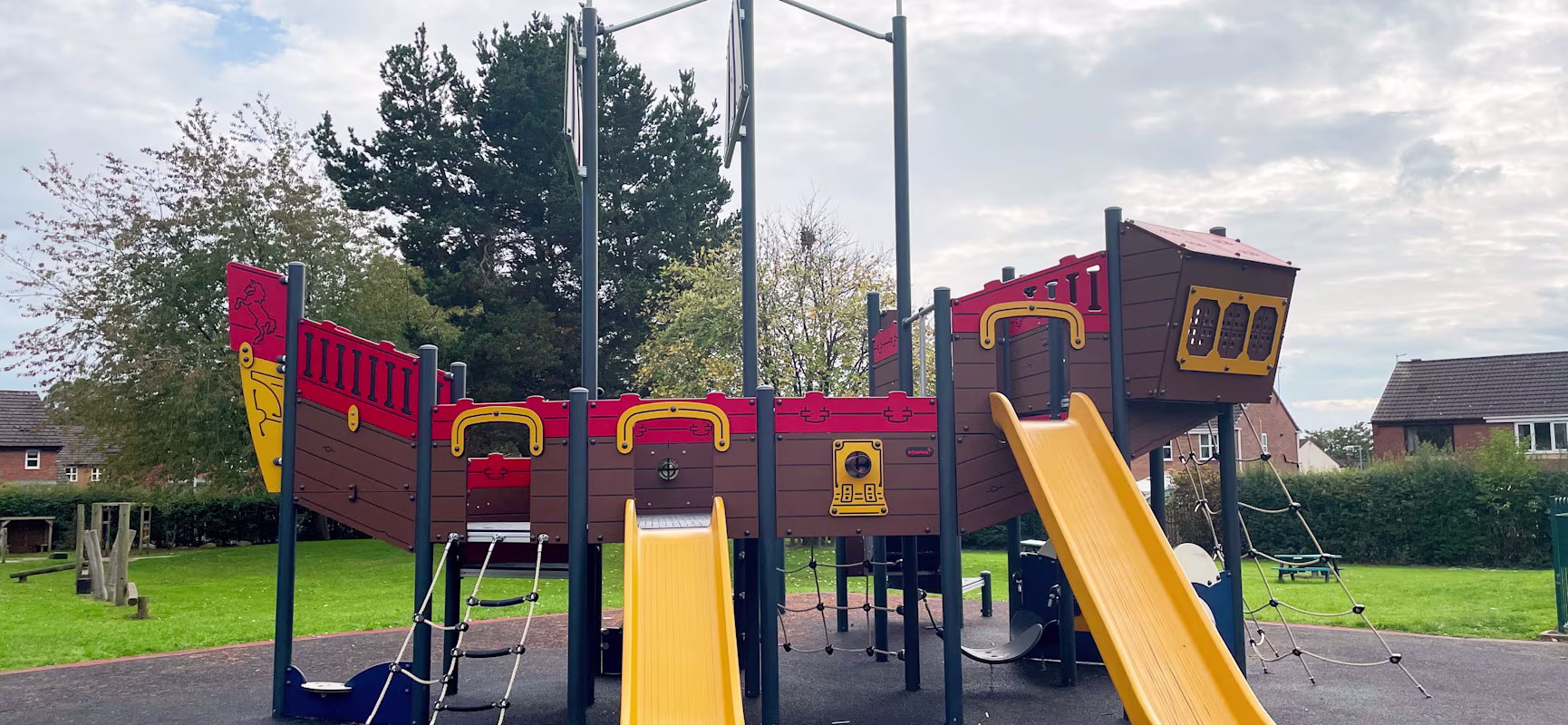 Colourful pirate ship playground with two bright yellow slides and climbing nets, set on soft ground in a green park with houses nearby