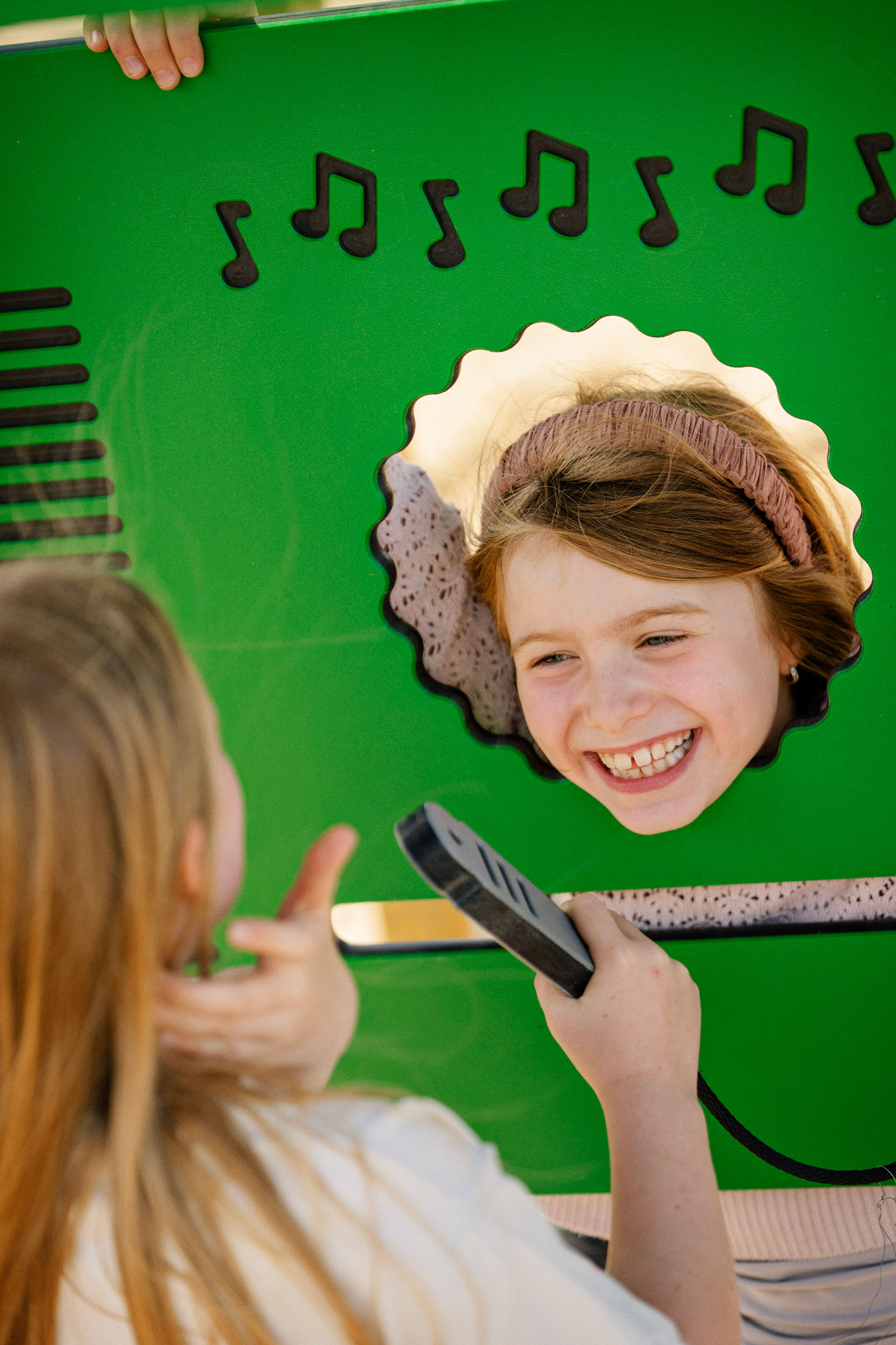 Child plays at a green musical panel, holding a toy microphone toward a friend whose face appears in a scalloped mirror opening.