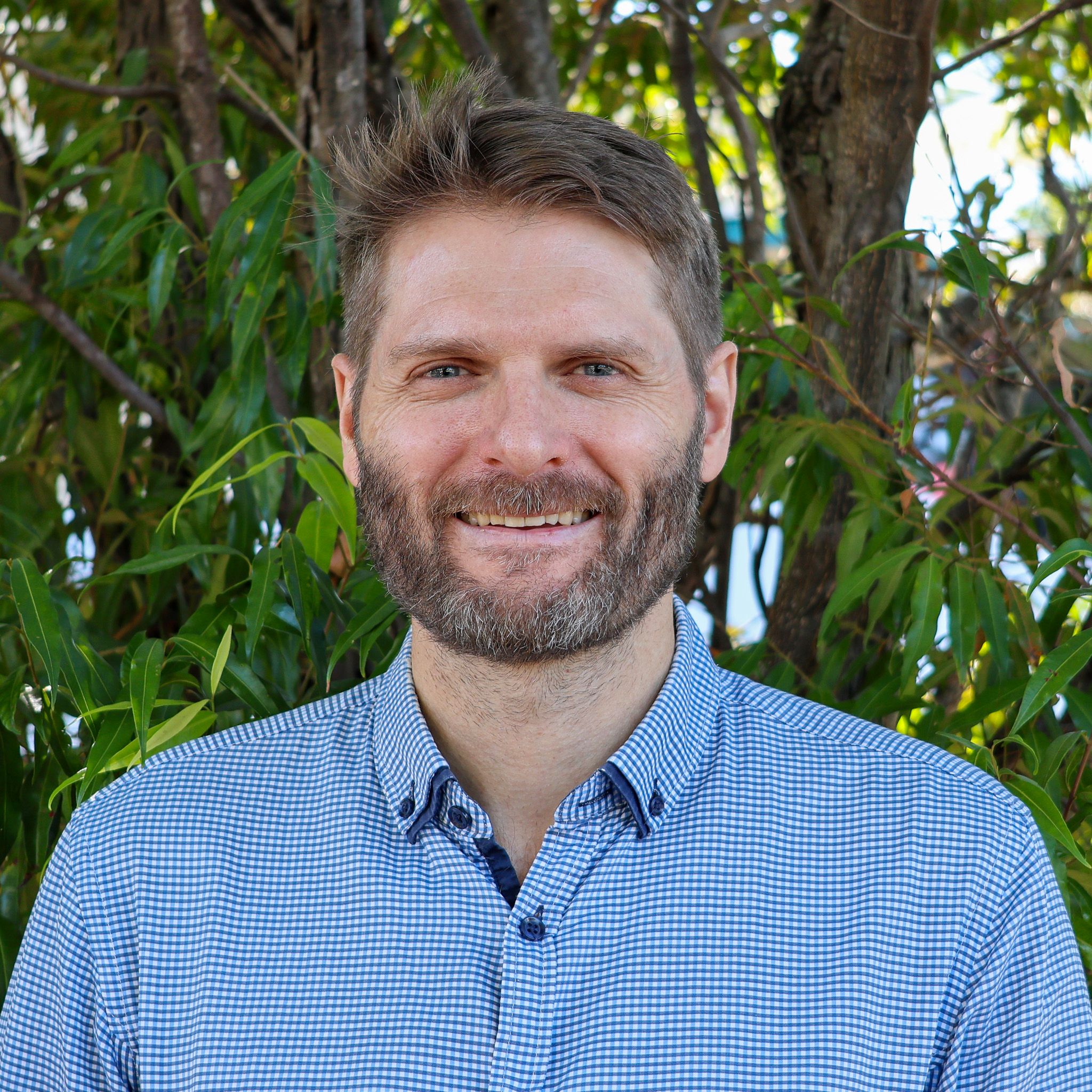 Person in a blue checkered shirt stands outdoors in front of green leafy trees, with their face blurred for privacy