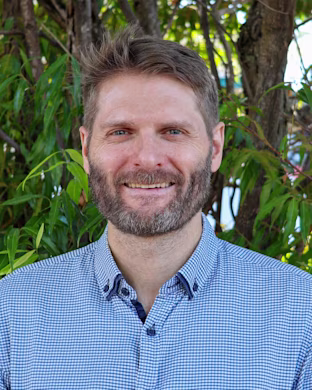Person in a blue checkered shirt stands outdoors in front of green leafy trees, with their face blurred for privacy