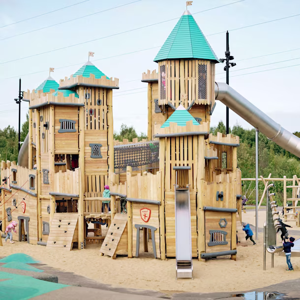 Large wooden playground castle sitting on top of sand surfacing. The castle has a large metal tunnel slide going from the top to the bottom. Showstopping pieces like this one are ideal for playground projects at zoos and other leisure facilities.