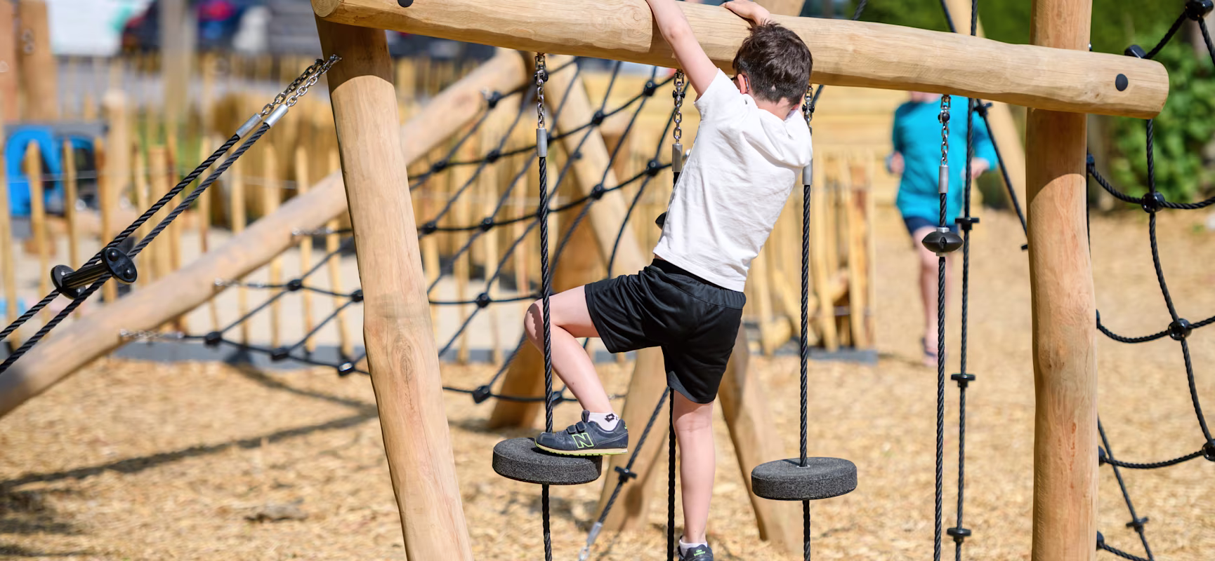 Child in white shirt and black shorts climbing a rope and wood obstacle at an outdoor playground on a sunny day.