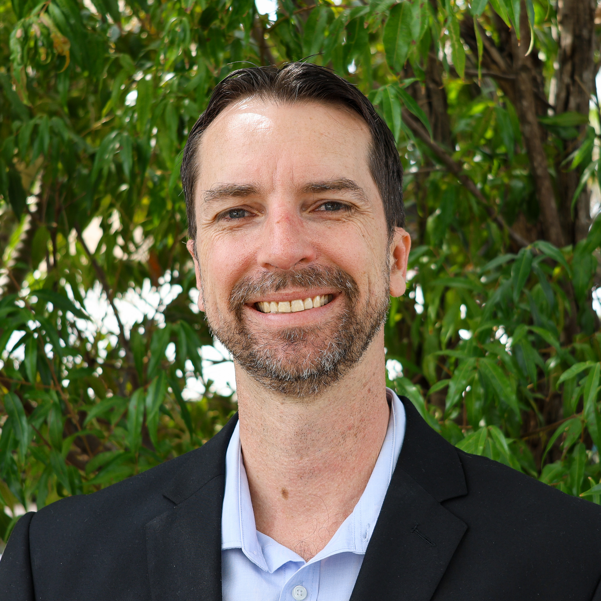 Person in a black blazer and light blue shirt standing outdoors in front of green leafy trees, with their face blurred.