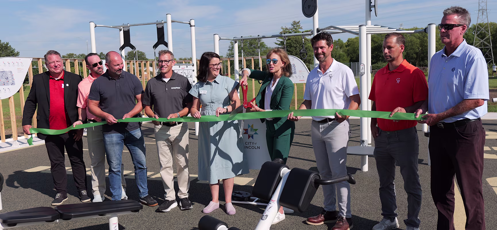 Group of officials cutting a long green ribbon at an outdoor fitness park opening on a sunny day, with exercise equipment behind them.