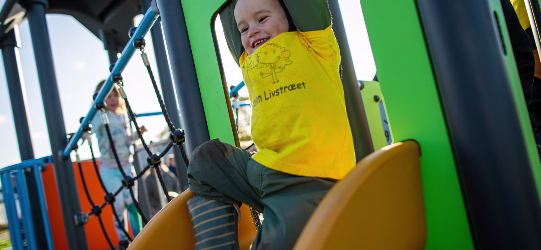 Child in yellow shirt and striped boots sliding down a yellow slide on a colourful playground, with another child on climbing ropes.