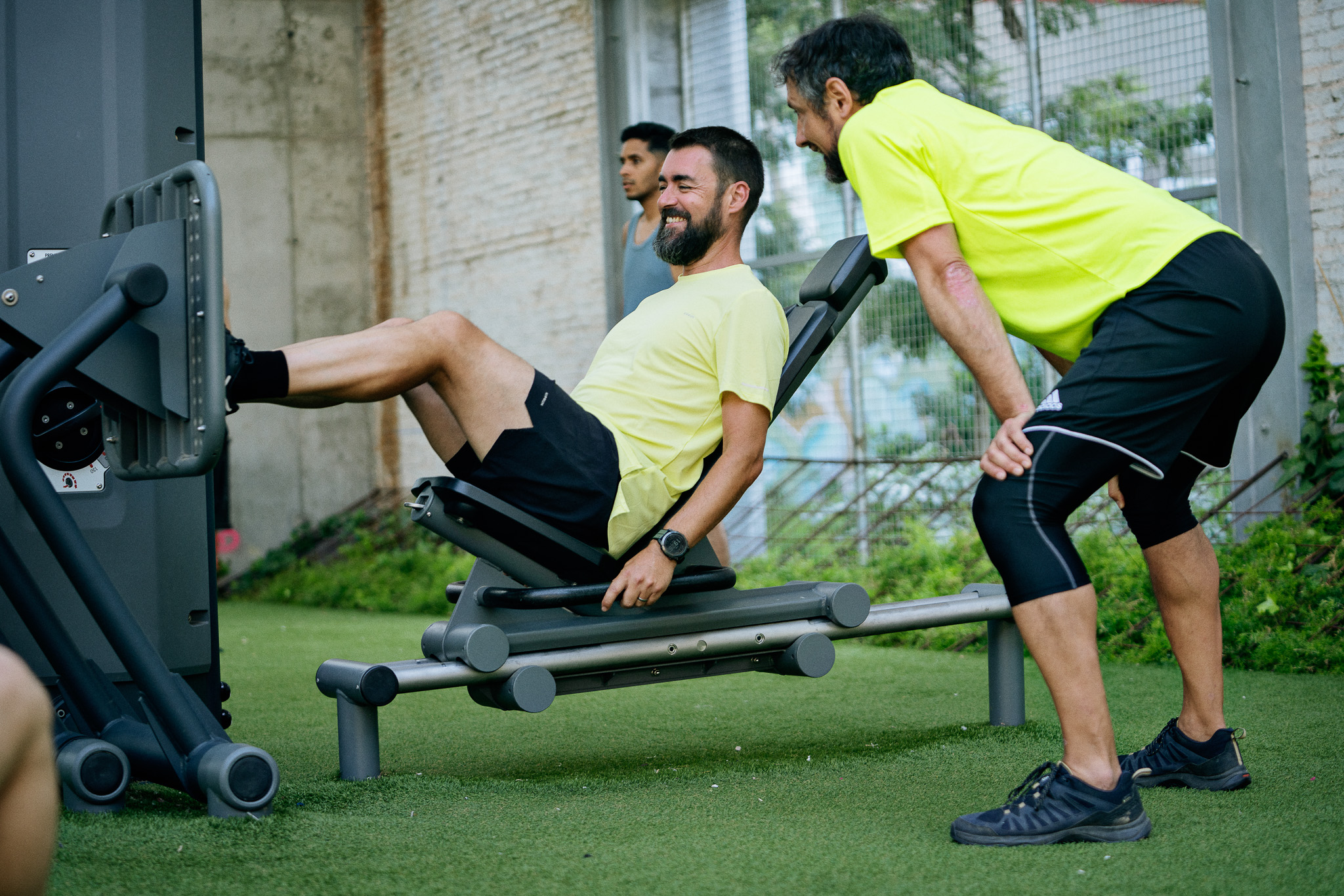 Man in yellow shirt using a leg press machine outdoors while a trainer in matching shirt and black shorts watches closely.