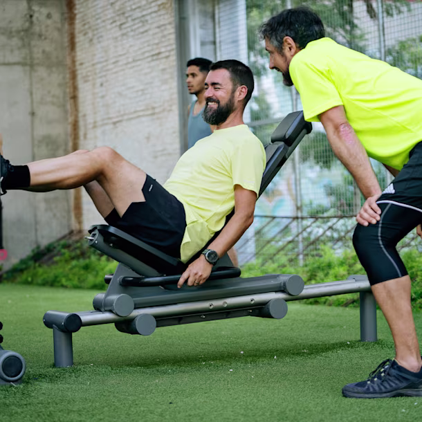 Man in yellow shirt using a leg press machine outdoors while a trainer in matching shirt and black shorts watches closely.