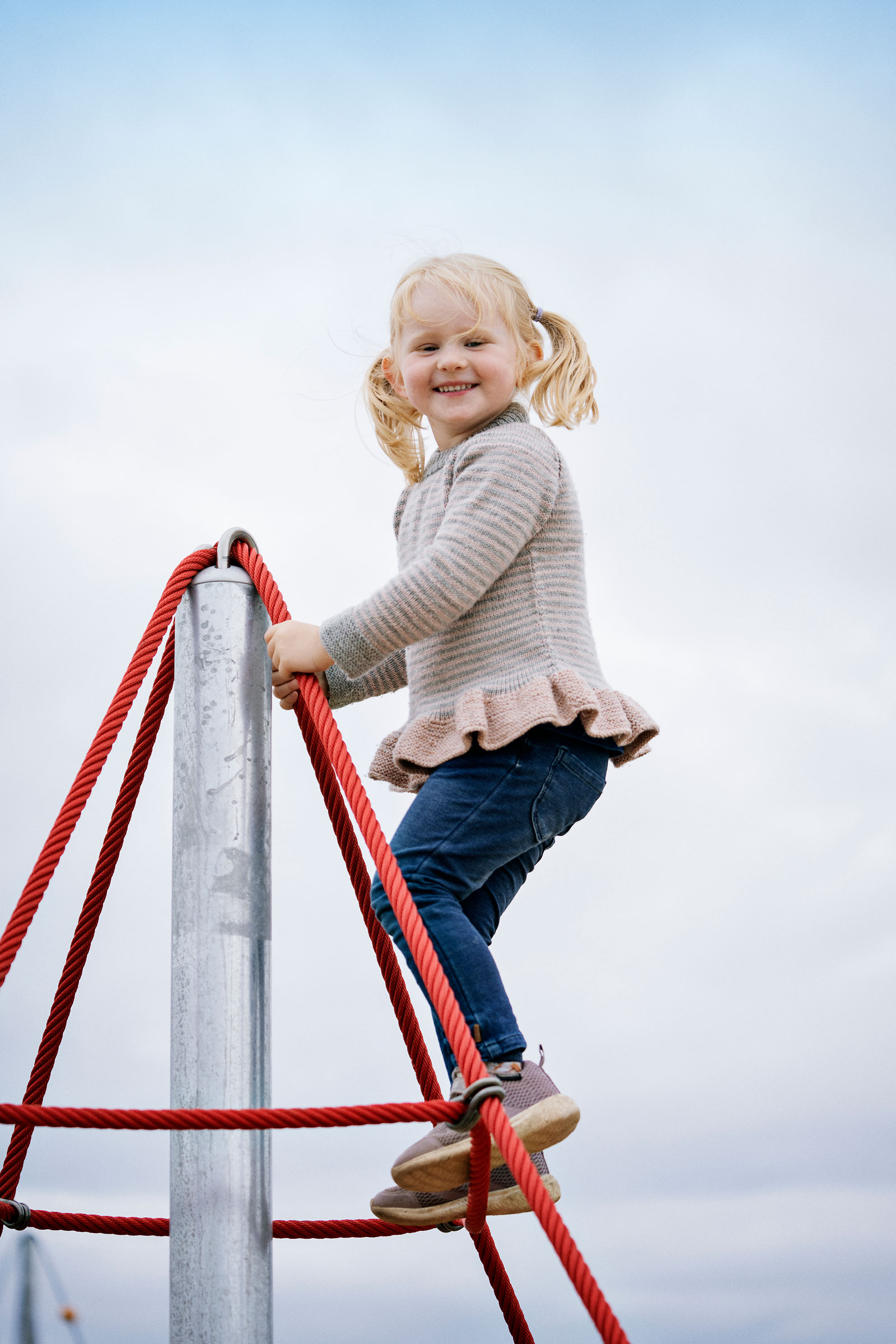 Child with pigtails climbing red rope playground structure, wearing striped sweater and jeans under a pale blue sky