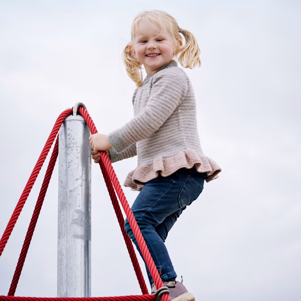 Child with pigtails climbing red rope playground structure, wearing striped sweater and jeans under a pale blue sky