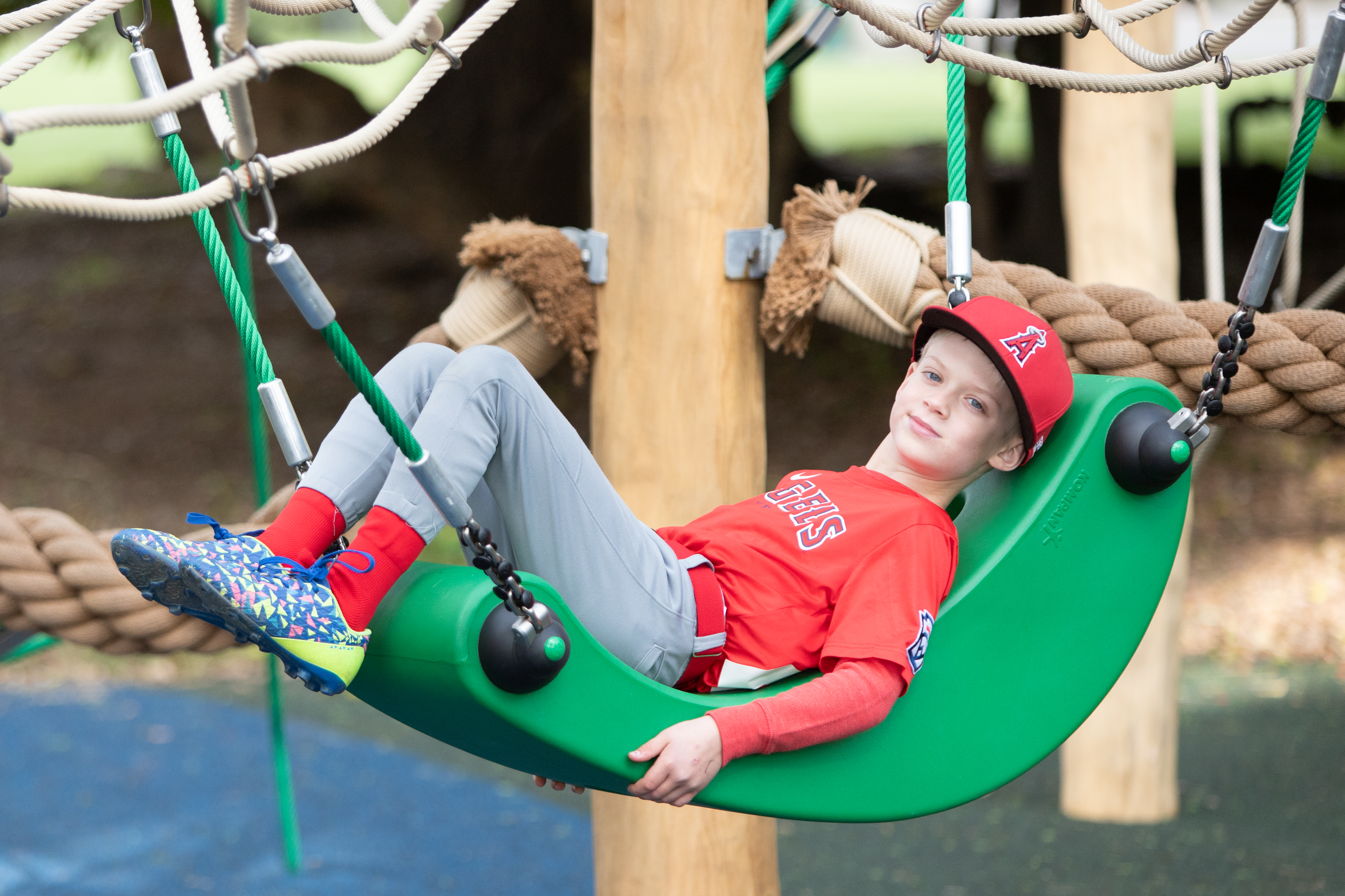 A child wearing a Los Angeles Angles baseball uniform smiling at the camera softly while relaxing on a hammock that's incorporated into a large, wooden commercial rope climbing structure