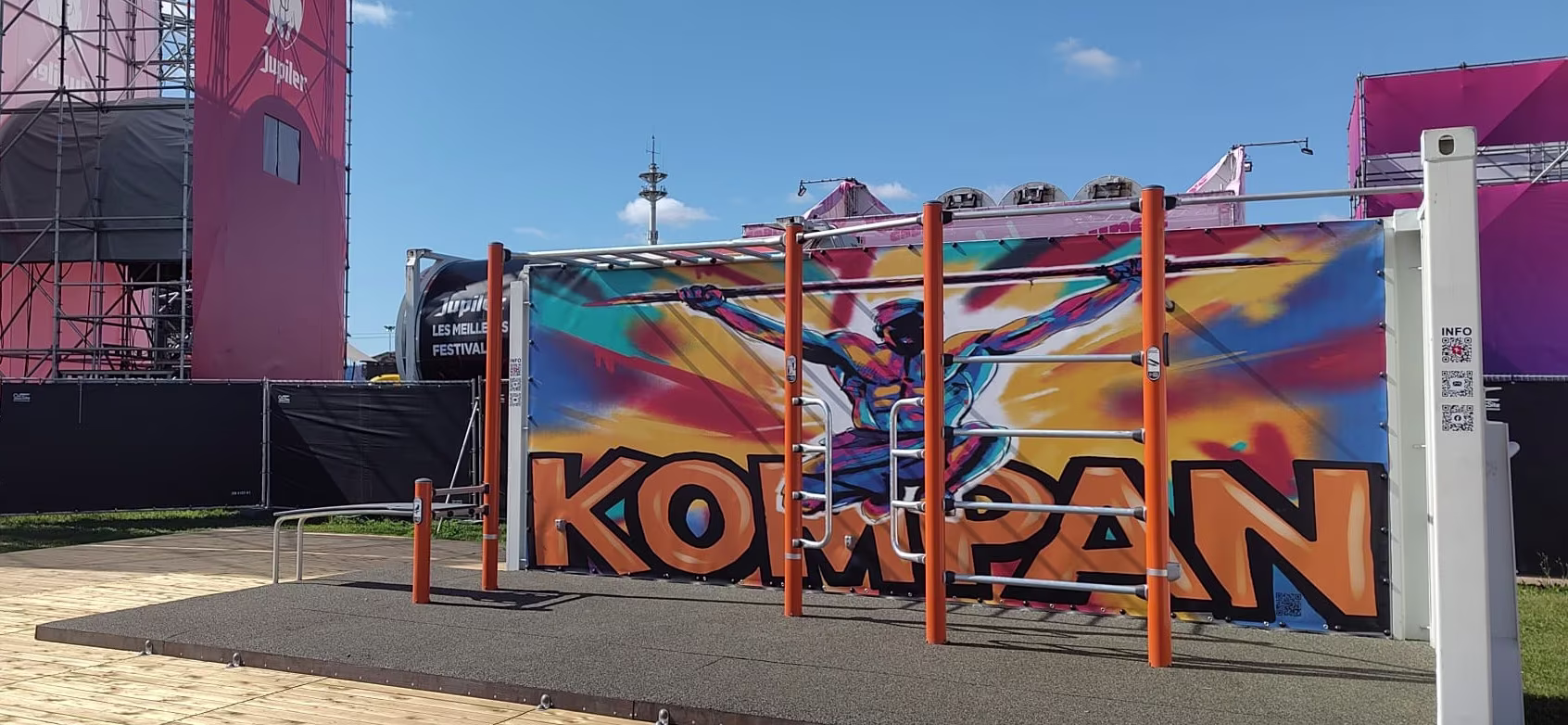 Outdoor fitness bars on a wooden deck in front of bright comic-style KOMPAN graffiti at a sunny festival site