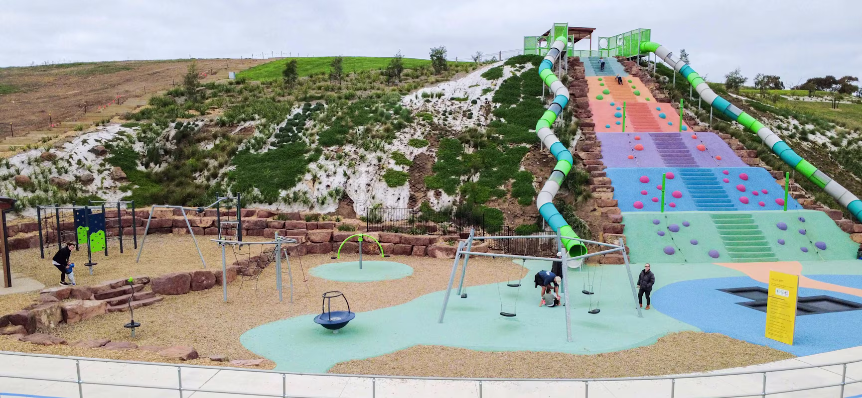 Colourful hillside playground with long tube slides, climbing zones, swings and families playing on soft-fall surfaces under cloudy sky