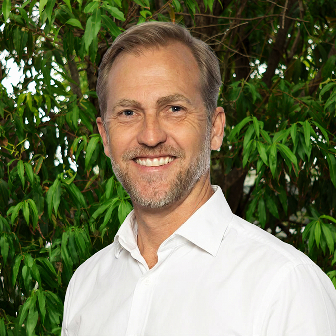 Person in a white shirt standing outdoors in front of lush green trees, with their face blurred for privacy