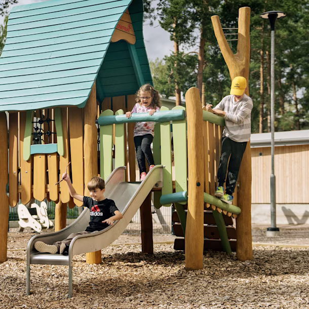 Children playing on a wooden playground structure with slide and climbing frame in a sunny park with trees and buildings nearby