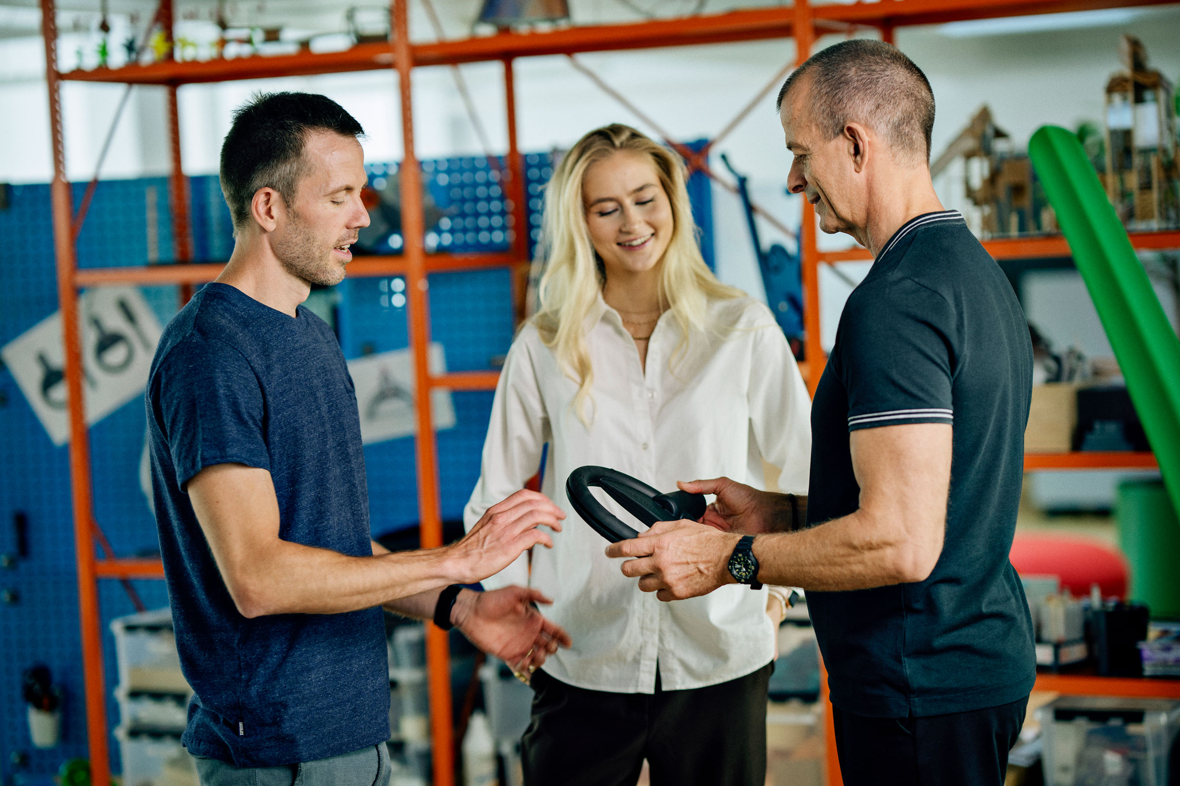 Three people stand in a workshop, discussing a black circular device one person is holding, with shelves and tools in the background.