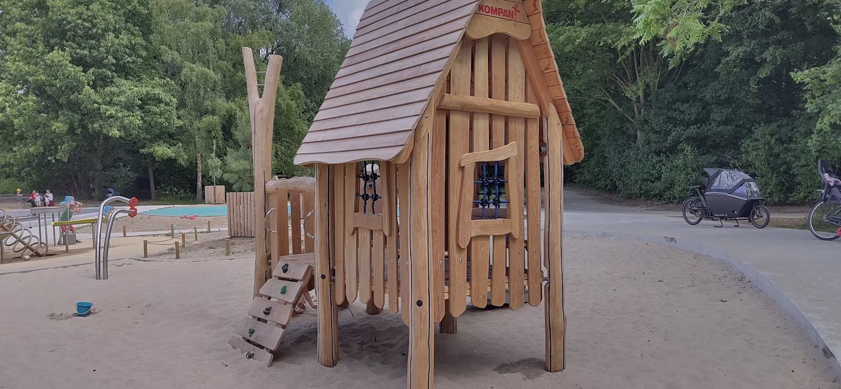 Wooden playhouse with ladder in a sandy playground, surrounded by trees, bikes and a water play area in the background.