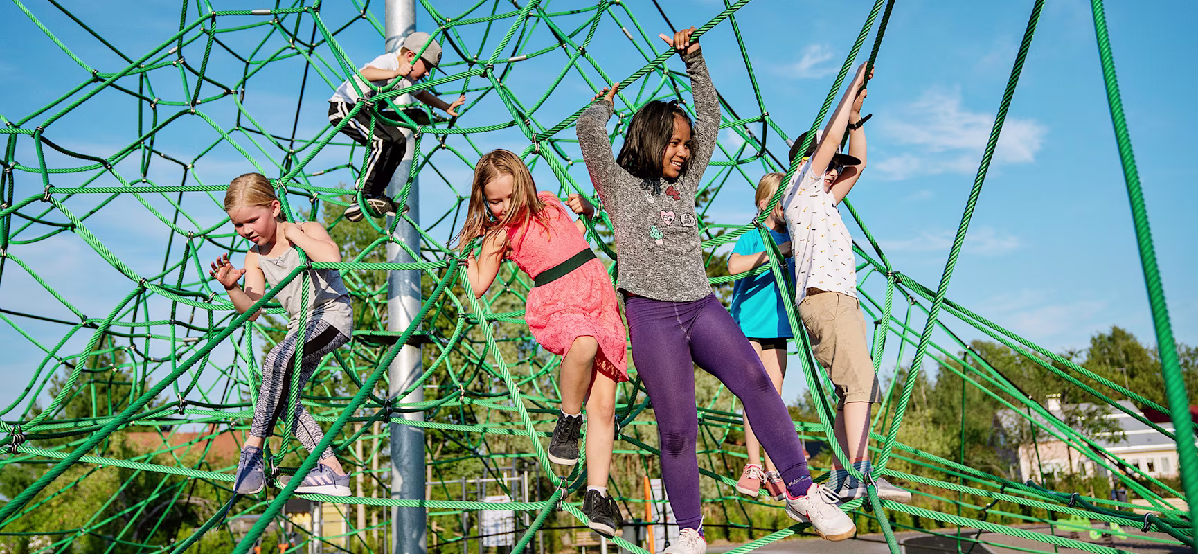 Children climbing and hanging on a large green rope climbing frame at an outdoor playground under a bright blue sky