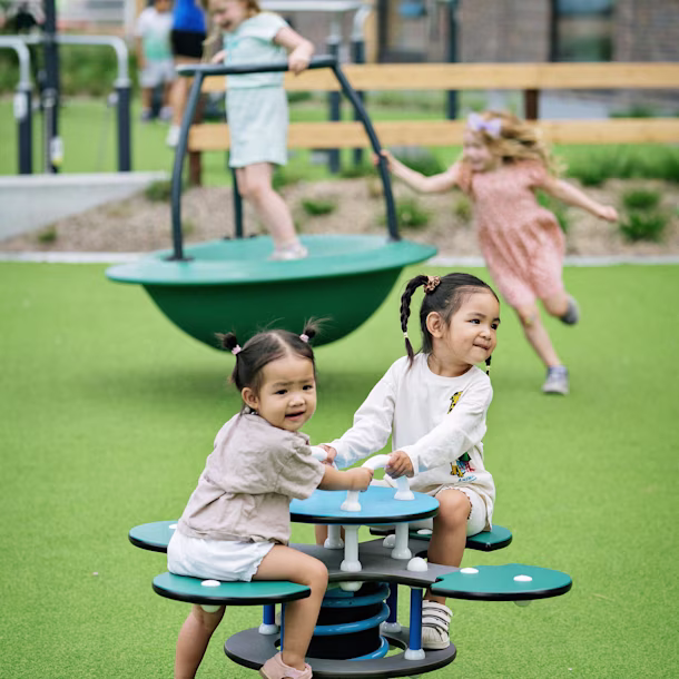 Two young girls playing on a green daisy springer with other young girls in the background on a tipi carousel. Creating zones within your playground layout plan can help younger children feel more confident to play.