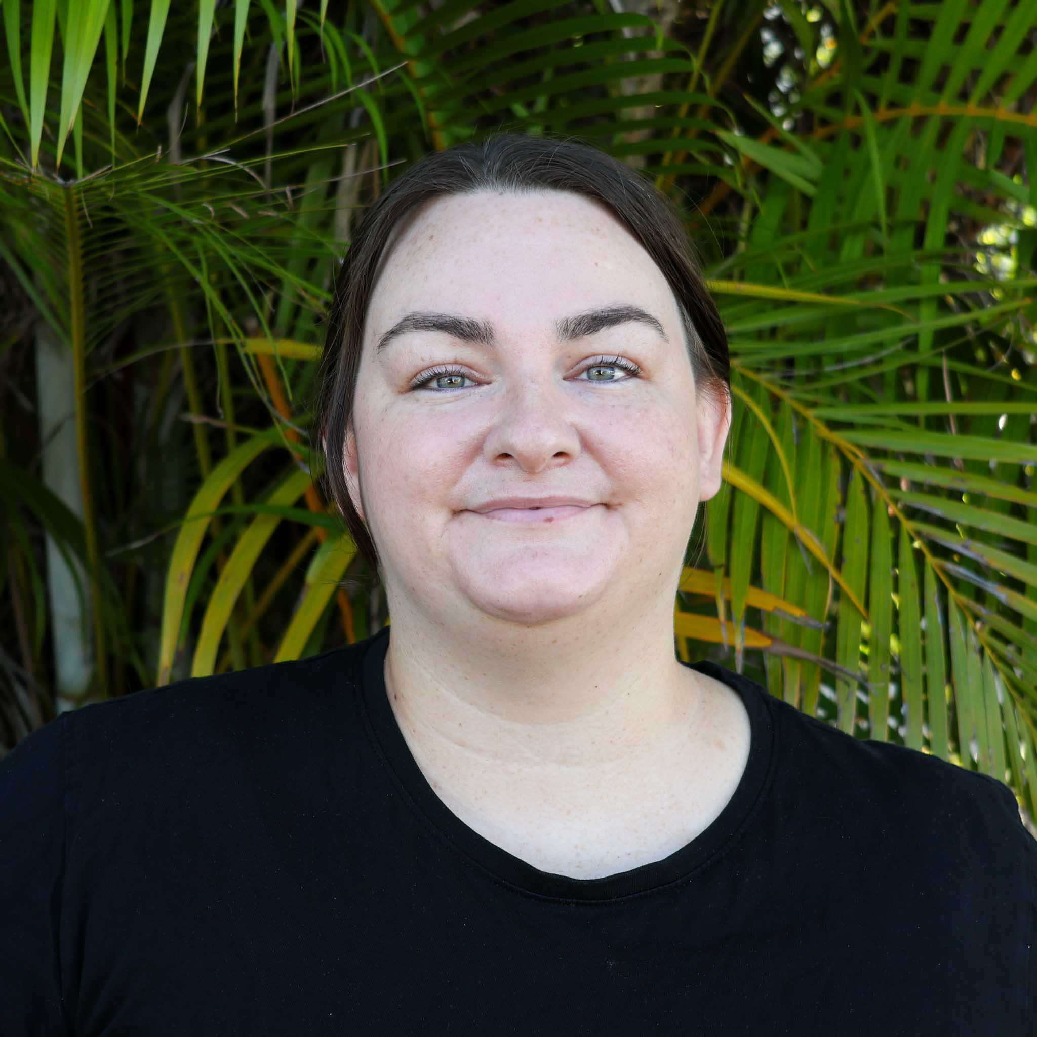 Person in a black shirt standing outdoors in front of dense green palm leaves, with their face obscured by a blurred square.