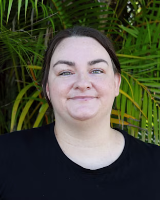 Person in a black shirt standing outdoors in front of dense green palm leaves, with their face obscured by a blurred square.