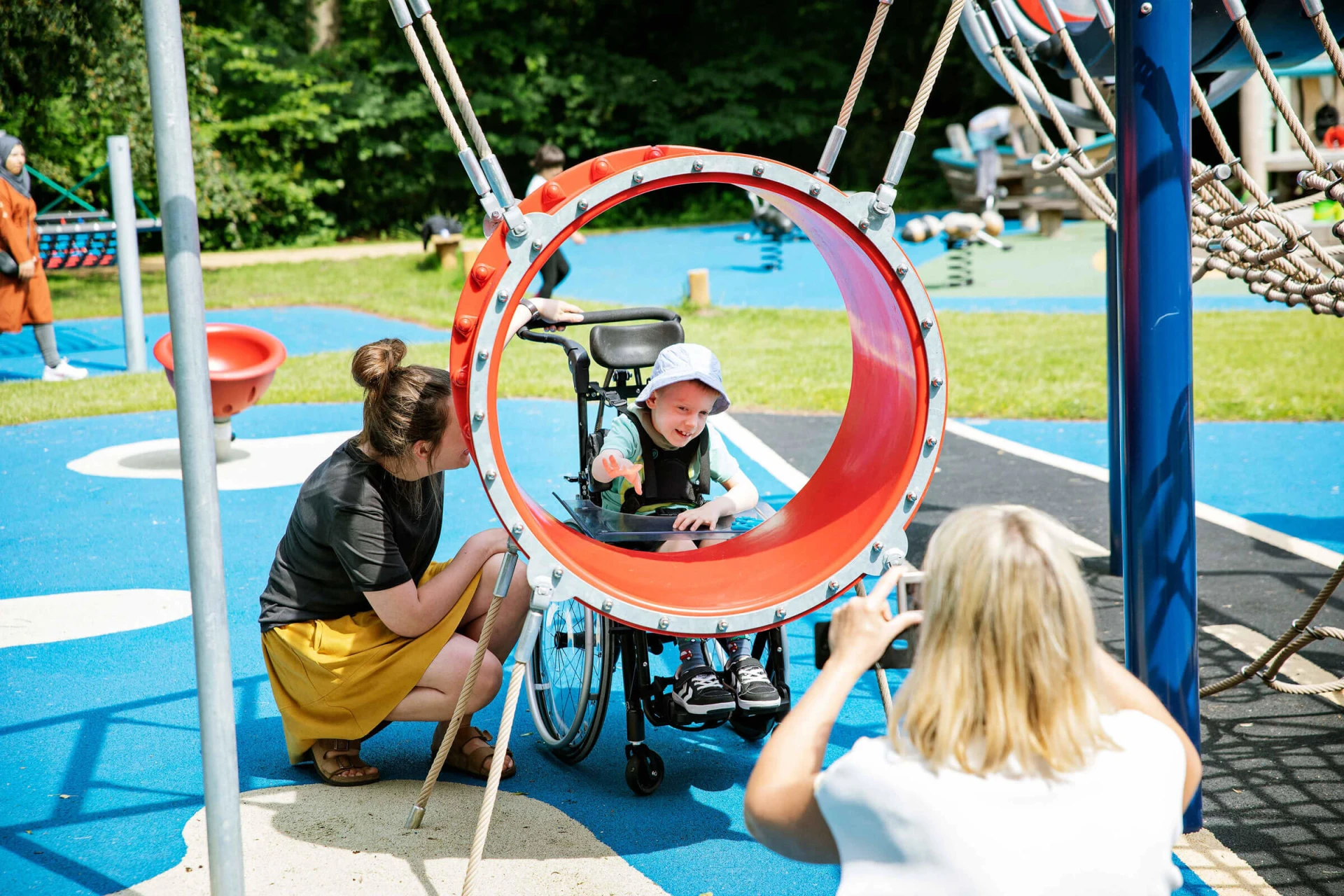 pyörätuolia käyttävä poika ja hänen hoitajiaan leikkimässä aistileikkivälineillä leikkipaikalla