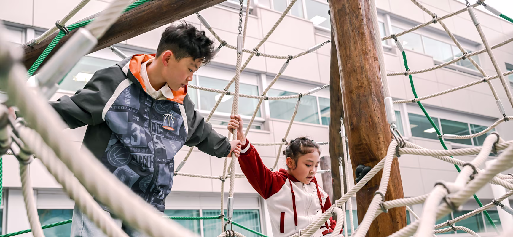 Two children on a rope climbing frame outdoors, holding on to nets and wooden poles in front of a modern building