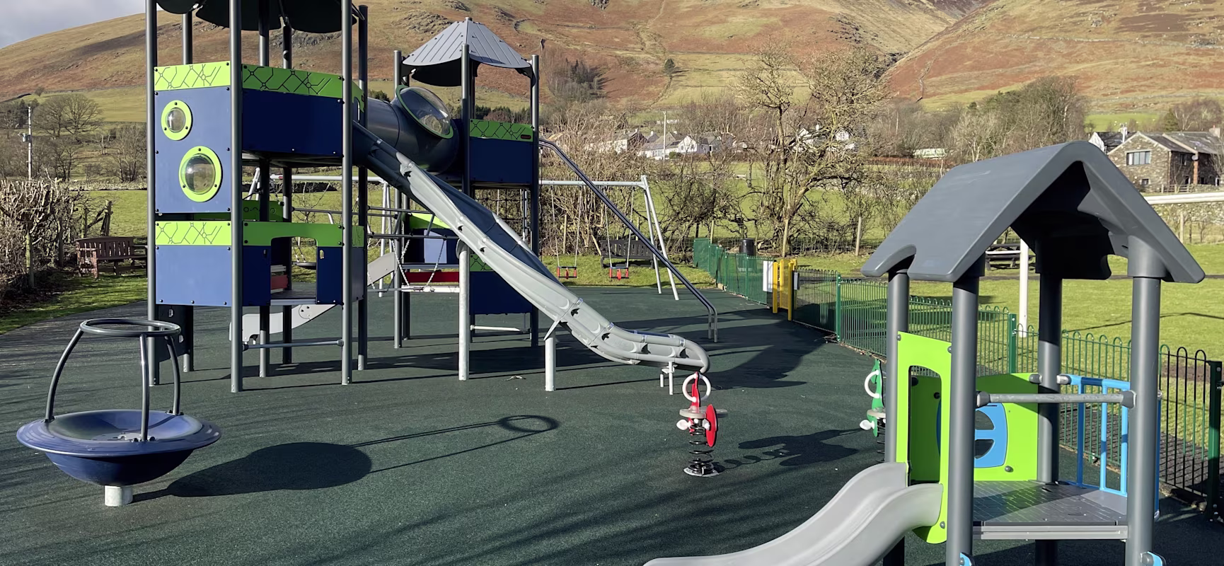 Modern playground with slides, climbing towers and spring riders on soft ground, set in a sunny rural park with hills in the background