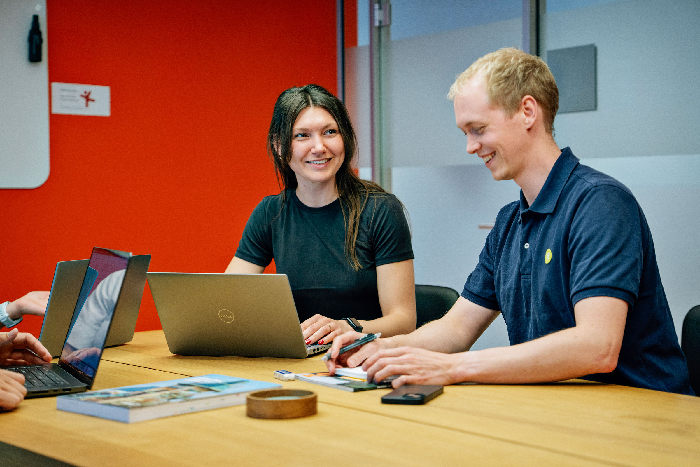 Two people with blurred faces sit at a table with laptops, notebooks, and phones, collaborating in a bright office with a red wall.