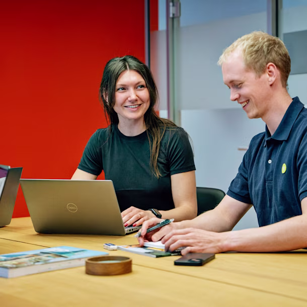 Two people with blurred faces sit at a table with laptops, notebooks, and phones, collaborating in a bright office with a red wall.