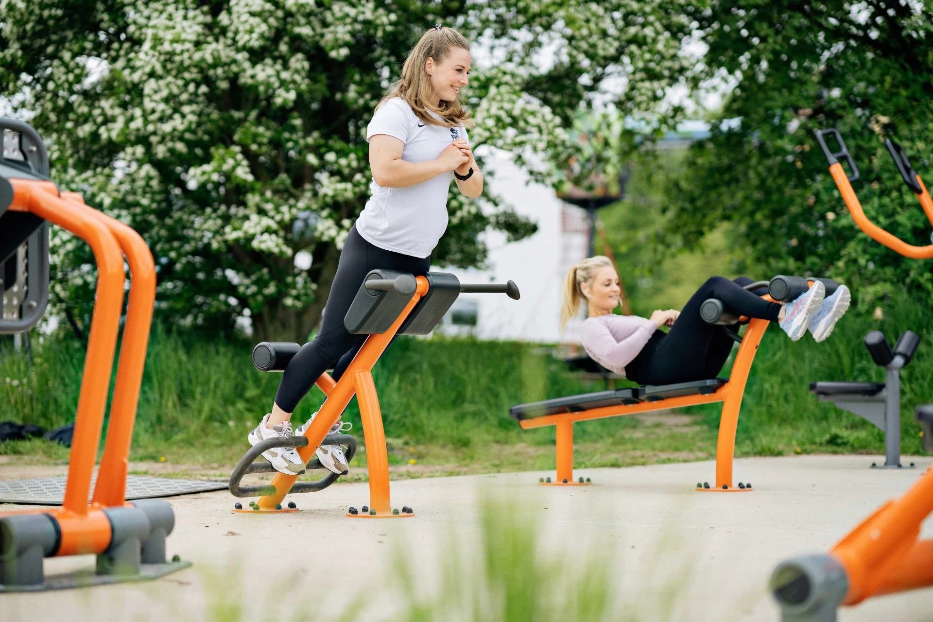 Two young women are exercising on bright orange outdoor gym equipment, surrounded by green trees. KOMPAN's playground planning tools and resources can help you create your next outdoor project. Browse our website to find out more.