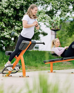 Two young women are exercising on bright orange outdoor gym equipment, surrounded by green trees. KOMPAN's playground planning tools and resources can help you create your next outdoor project. Browse our website to find out more.