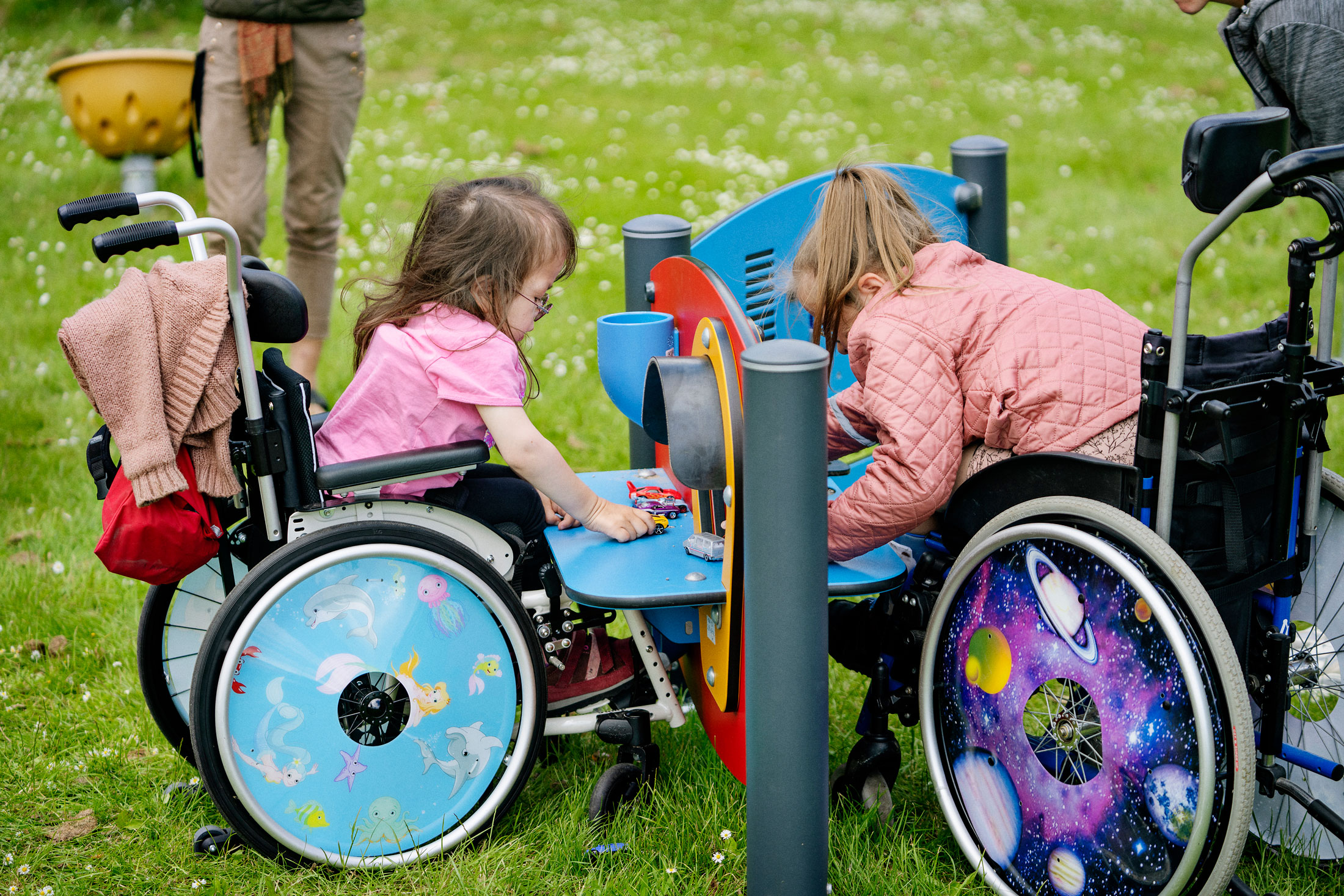Two children in wheelchairs with colourful wheel covers play together at an accessible outdoor play station on green grass.