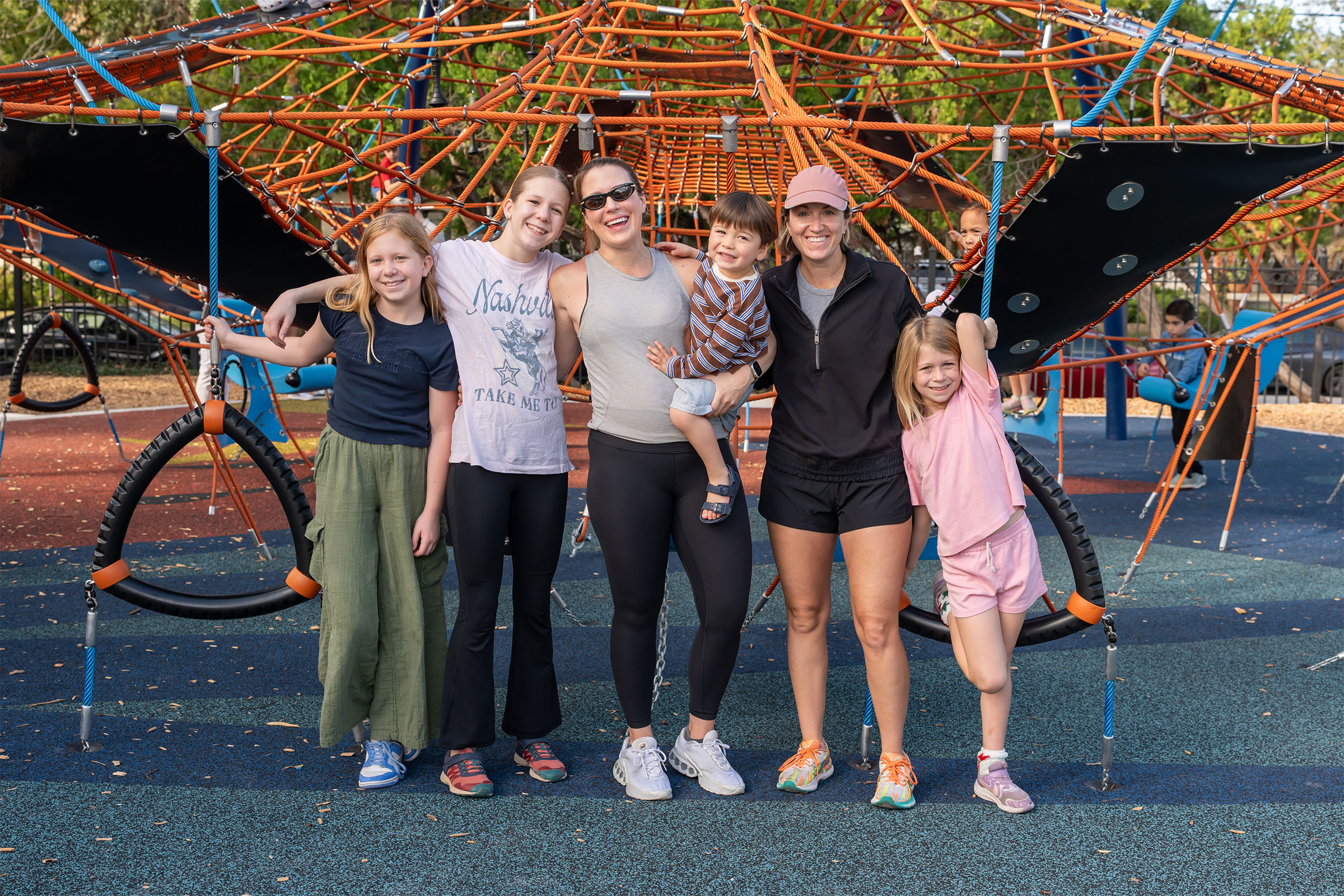Group of adults and children posing together at a colourful playground climbing frame on a sunny day