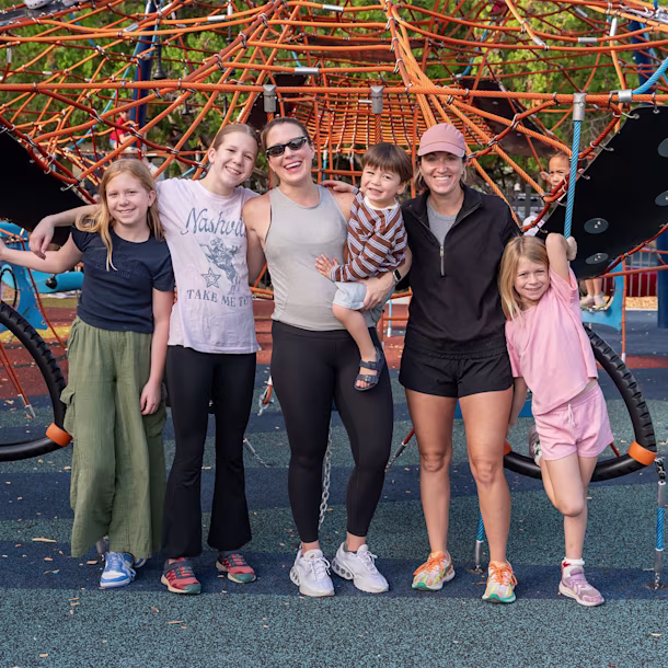 Group of adults and children posing together at a colourful playground climbing frame on a sunny day