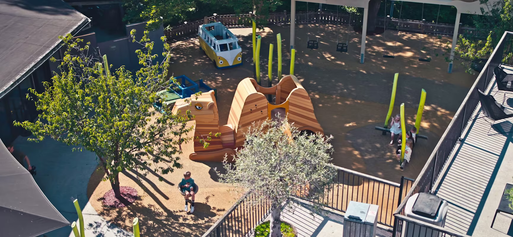 Aerial view of a sunny playground with camel and bus structures, swings, trees and children playing on green poles.