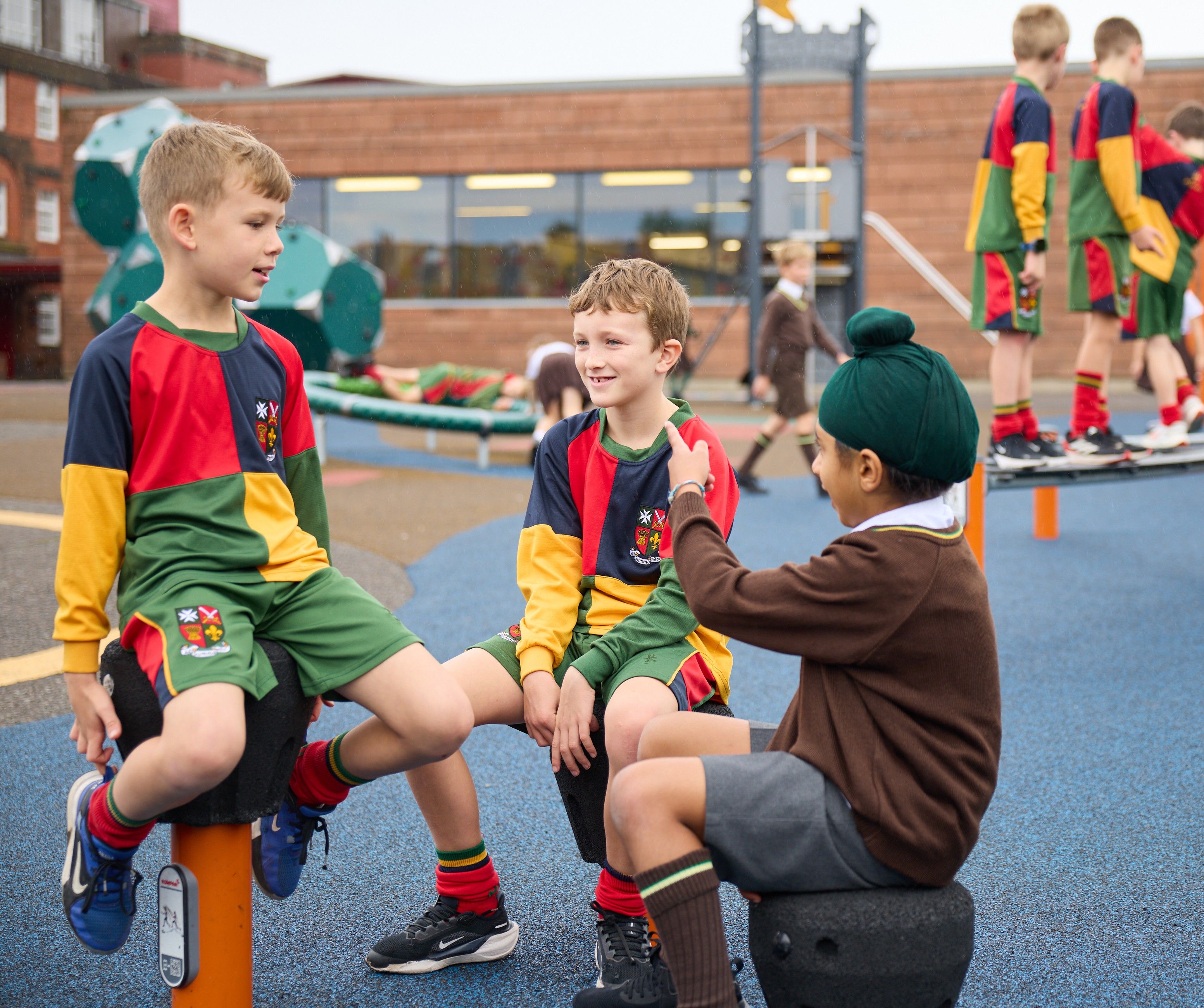 Children talk and sit on playground stools; two in colorful sports kits, one in school uniform, with others playing in background