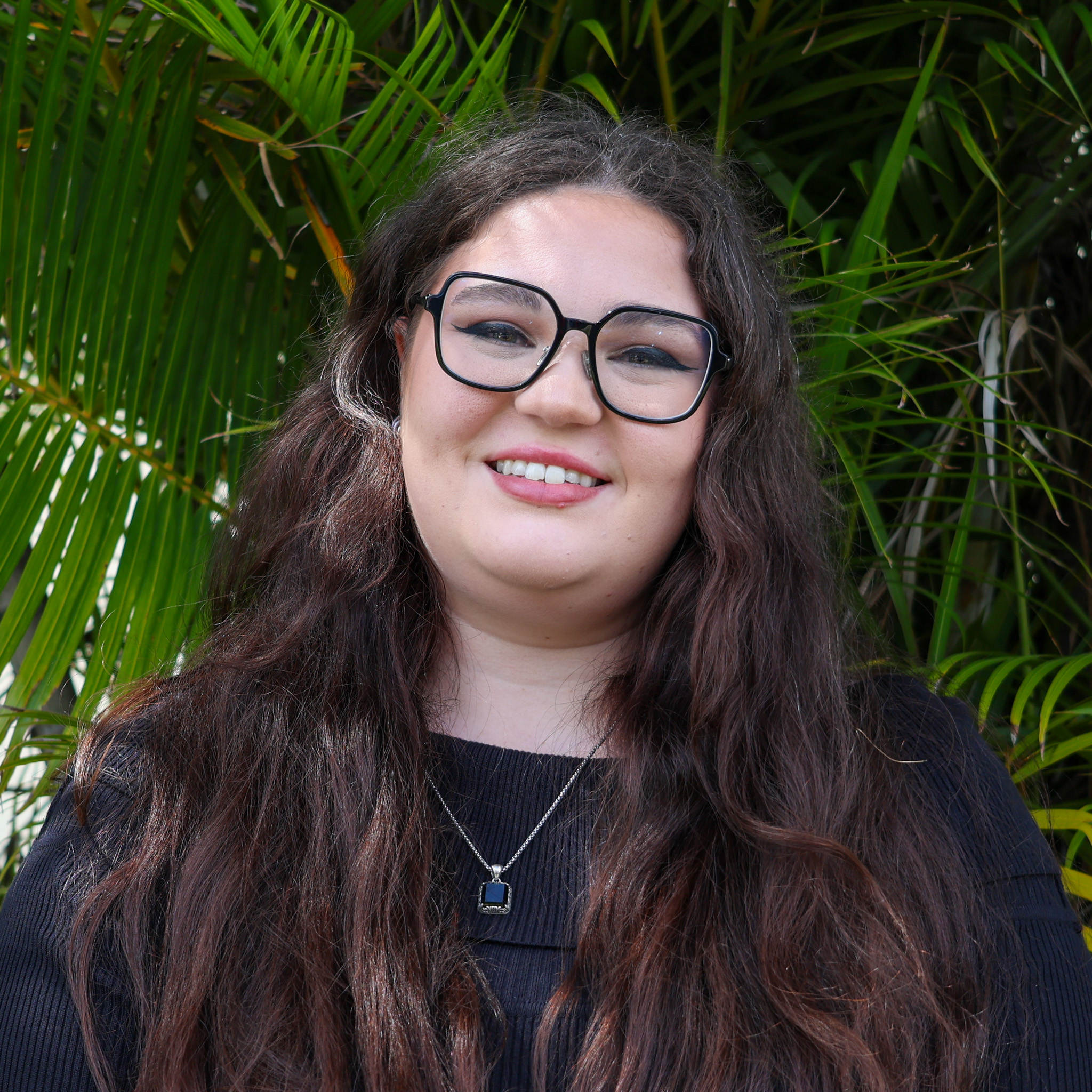 Person with long wavy brown hair in a dark top and pendant necklace, standing in front of green palm leaves outdoors