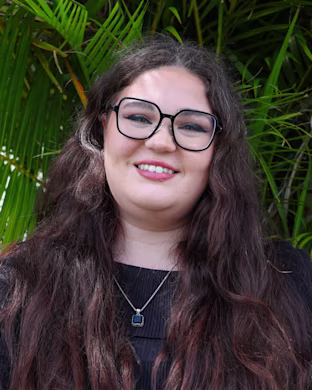 Person with long wavy brown hair in a dark top and pendant necklace, standing in front of green palm leaves outdoors
