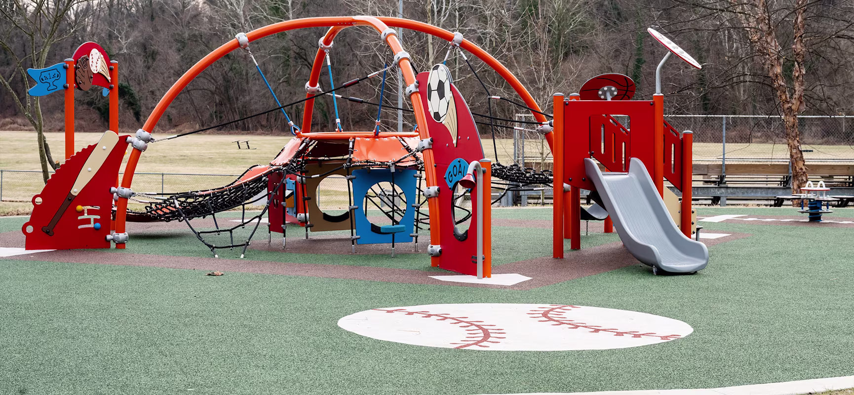 Colorful sports-themed playground with climbing nets, slides, and arches on rubber turf, set in a park with trees and open field.