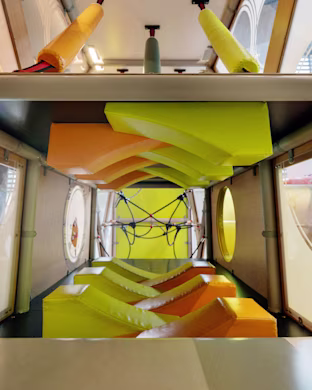 Interior of a bright indoor play tunnel with yellow and orange foam obstacles and climbing ropes at the far end