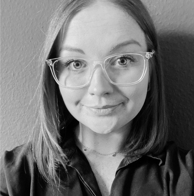 Black-and-white portrait of a person with shoulder-length hair and glasses, wearing a dark shirt and delicate layered necklaces.