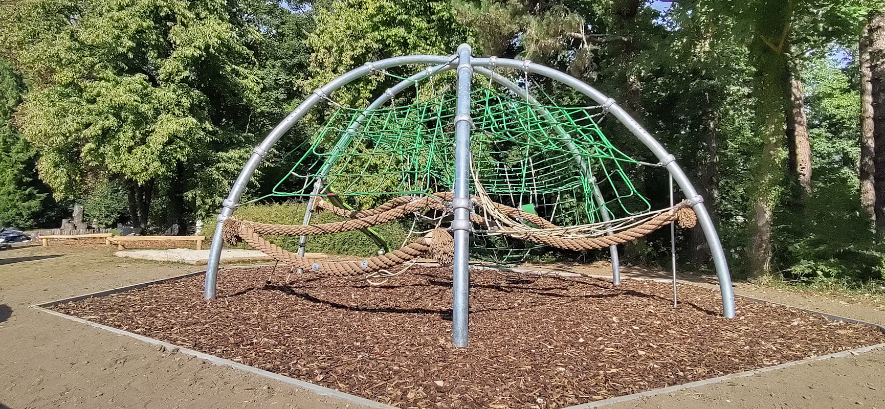 Metal-framed playground climbing dome with green nets and rope hammocks on woodchip ground, set in a sunny park with tall trees