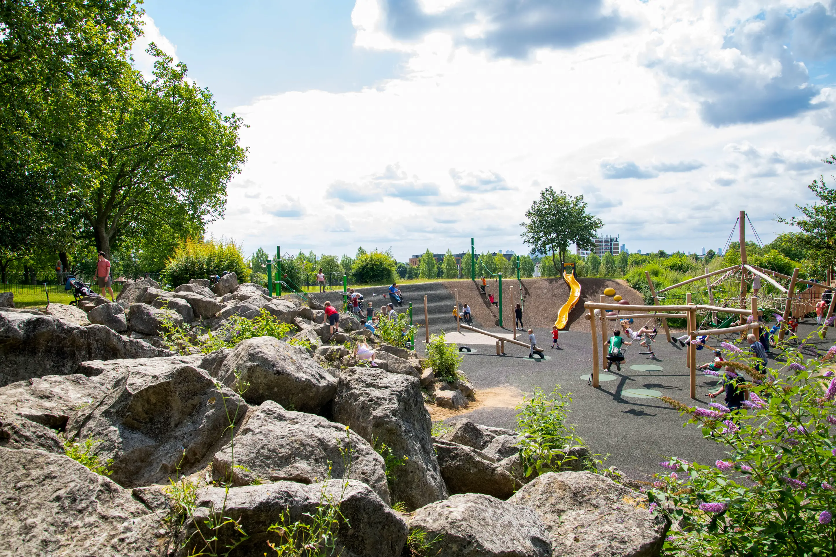 Natural adventure playground with wooden climbing structures, rope bridges, a metal slide, and steps set among lush green trees and grass