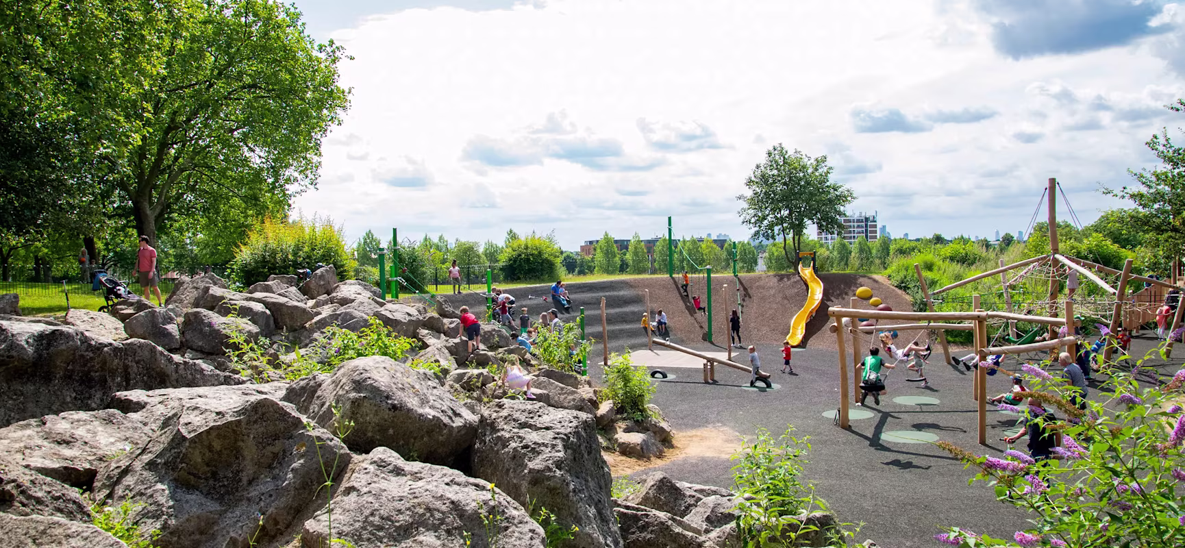 Natural adventure playground with wooden climbing structures, rope bridges, a metal slide, and steps set among lush green trees and grass