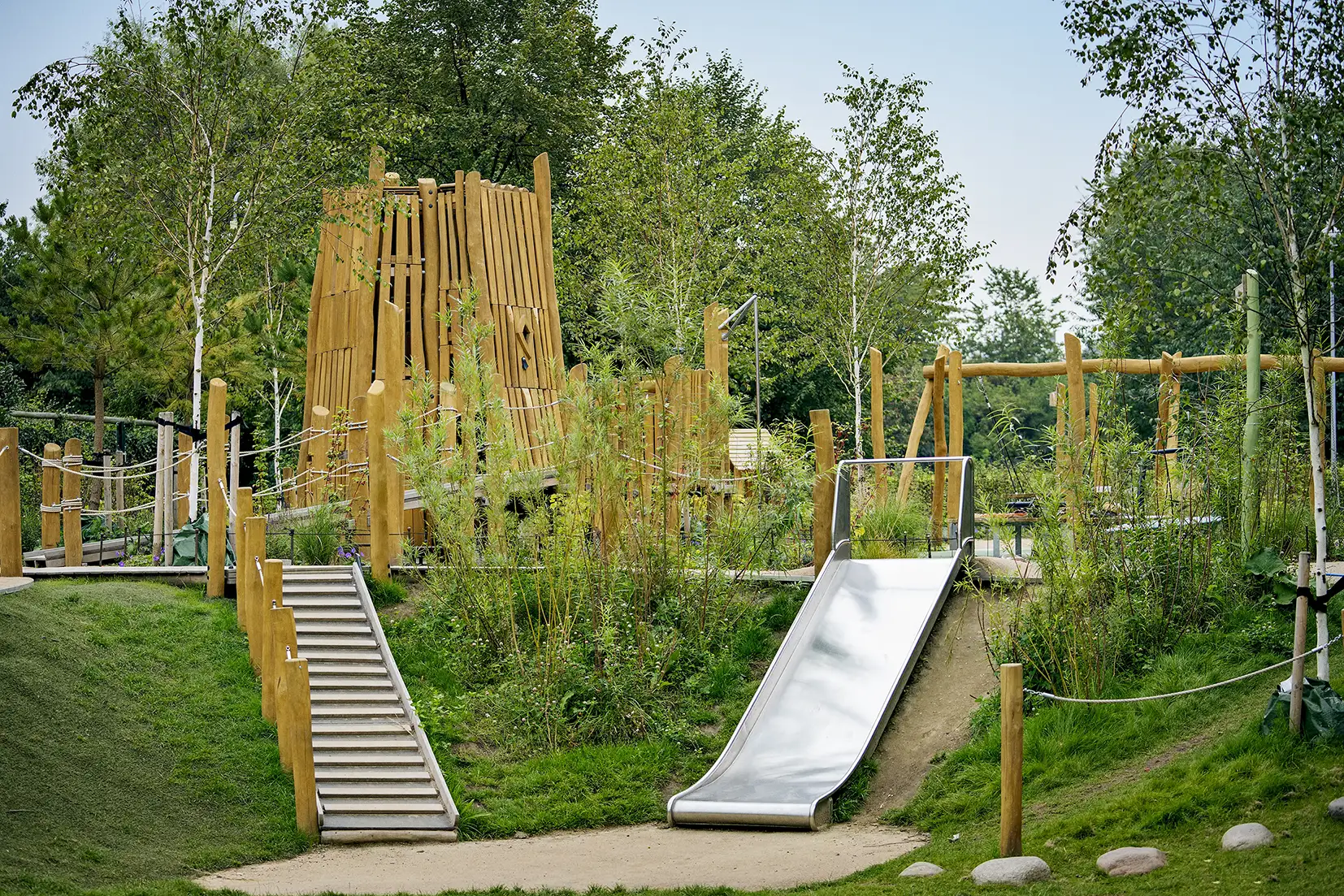 Natural adventure playground with wooden climbing structures, rope bridges, a metal slide, and steps set among lush green trees and grass