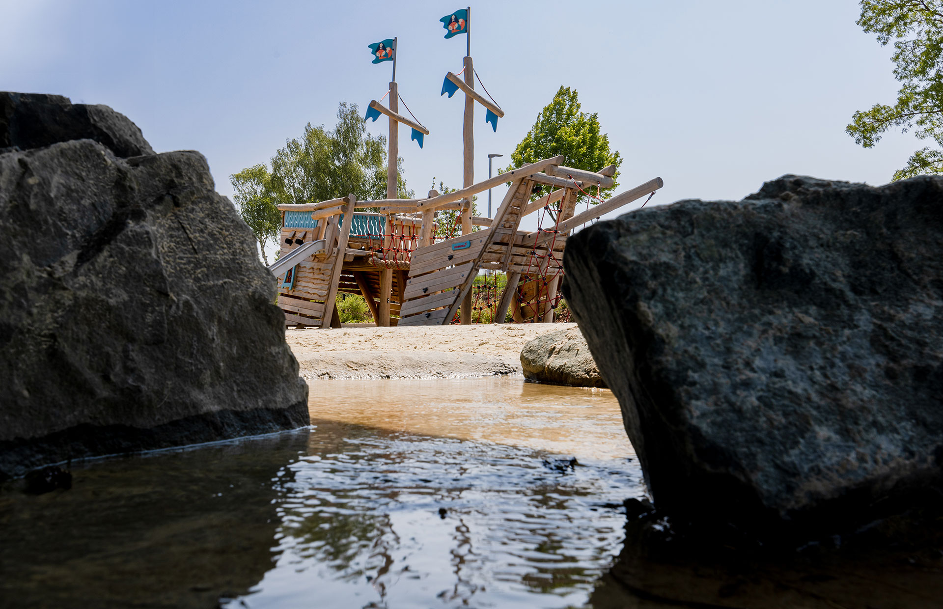 Wooden pirate ship playground with blue flags, seen between large rocks by a shallow stream on a sunny day.