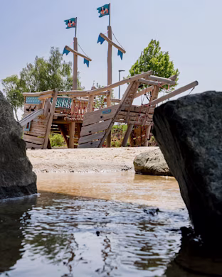 Wooden pirate ship playground with blue flags, seen between large rocks by a shallow stream on a sunny day.