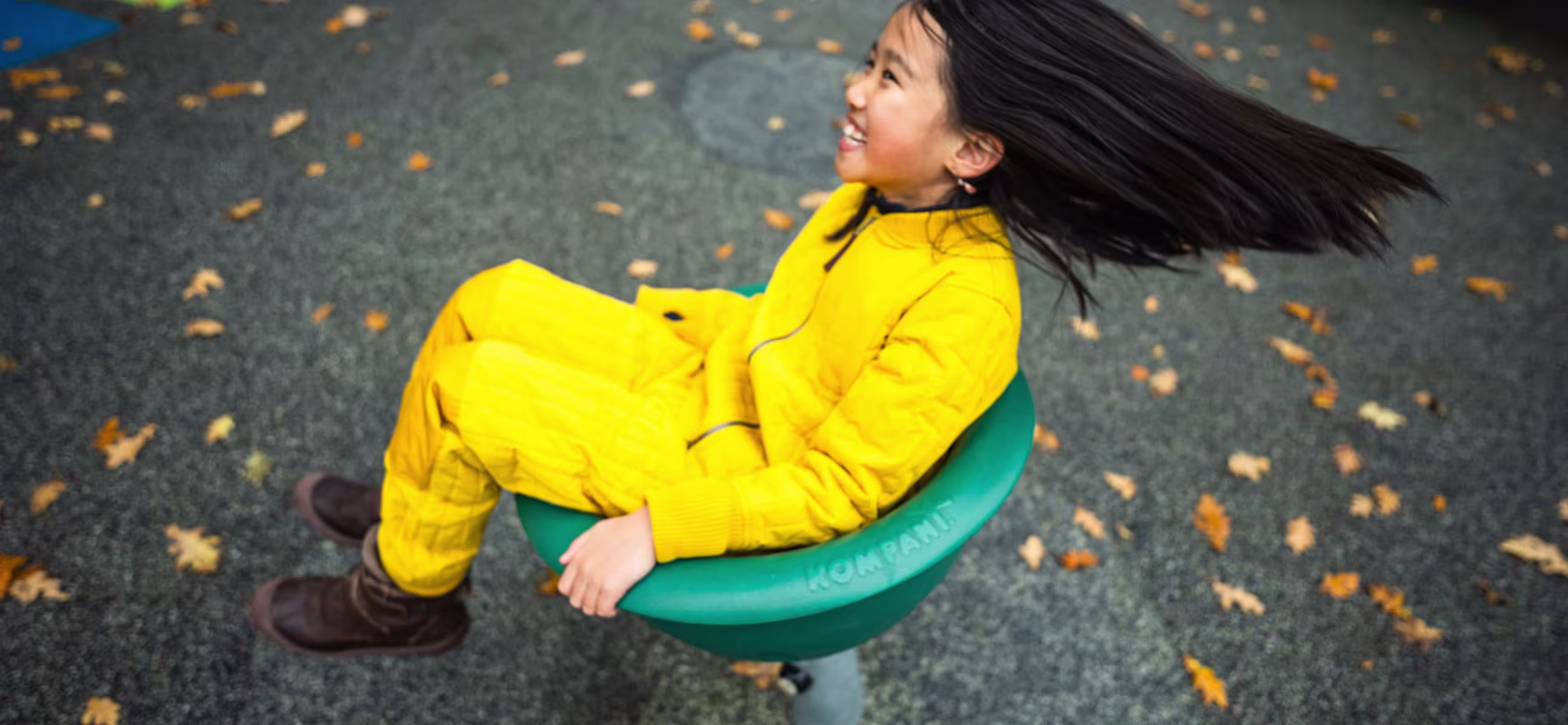 Child in yellow outfit spinning on a green playground seat, hair flying, with scattered autumn leaves on rubber ground