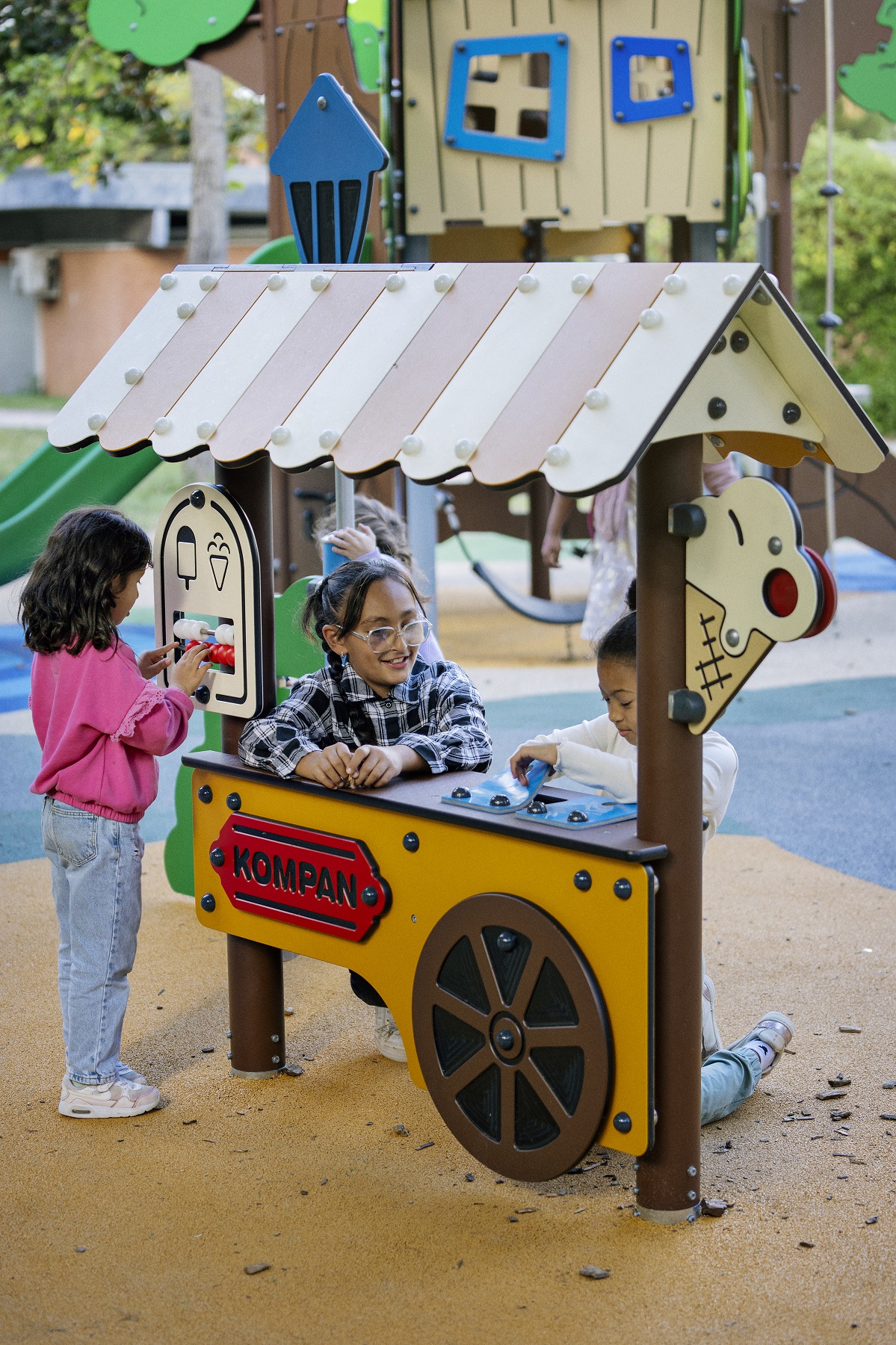 Children play at a colorful Kompan play stand in a park, pretending to sell ice cream under a small toy house roof.